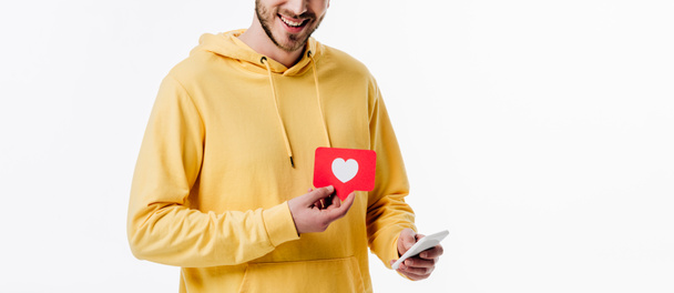 panoramatický záběr mladého muže ve žlutém mikina přidržený smartphone a červený papír s kartou s symbolem srdce izolovaným na bílém - Fotografie, Obrázek