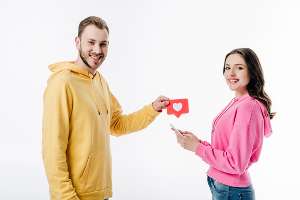 smiling handsome man giving red paper cut card with heart symbol to pretty girl using smartphone isolated on white - Φωτογραφία, εικόνα