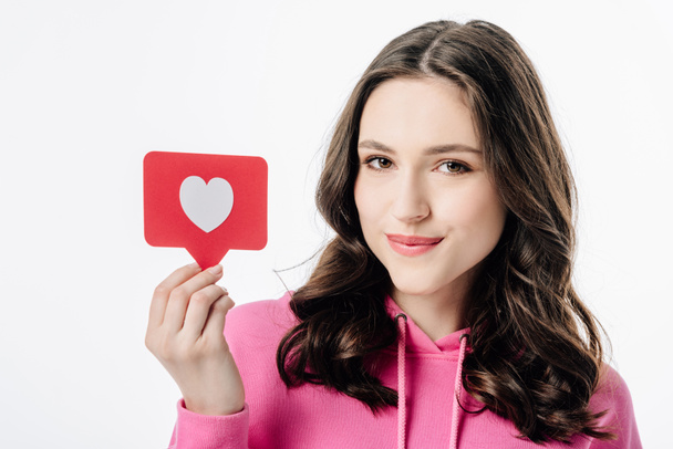 pěkná usměvavá dívka s červeným papírem, s symbolem srdce a s pohledem na kameru izolovanou na bílém - Fotografie, Obrázek