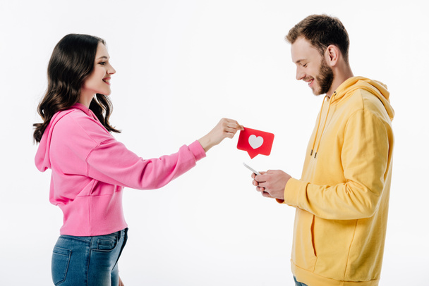 side view of pretty girl giving red paper cut card with heart symbol to boyfriend using smartphone isolated on white - Foto, immagini