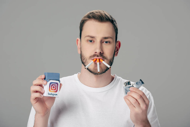 young man with cigarettes in mouth holding pack with instagram logo and looking at camera isolated on grey - Photo, image