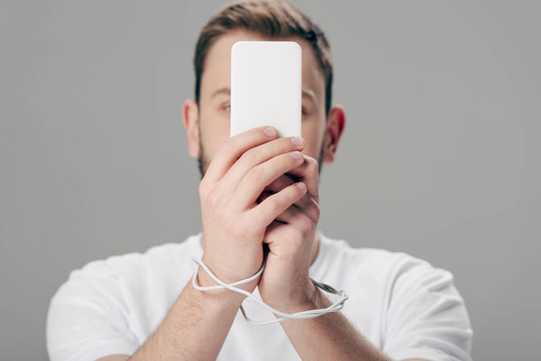 giovane uomo con cavo usb intorno alle mani utilizzando smartphone isolato su grigio
 - Foto, immagini