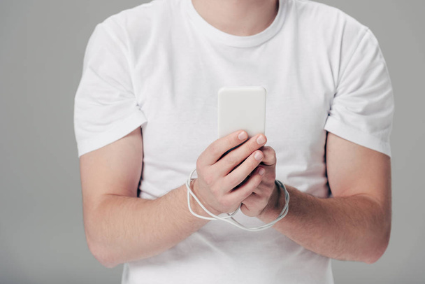 灰色で隔離されたスマートフォンを使用して手の周りにUSBケーブルを持つ若者の部分的なビュー - 写真・画像
