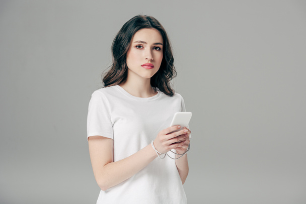 attrayant jeune femme en t-shirt blanc et câble USB autour des mains en utilisant smartphone isolé sur gris
 - Photo, image