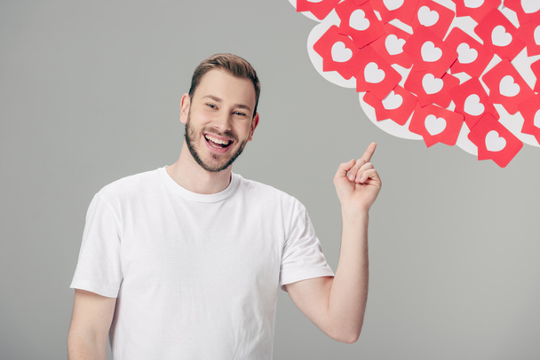 vrolijke jonge man in wit t-shirt wijzend met vinger op rood papier gesneden kaarten met harten symbolen geïsoleerd op grijs - Foto, afbeelding