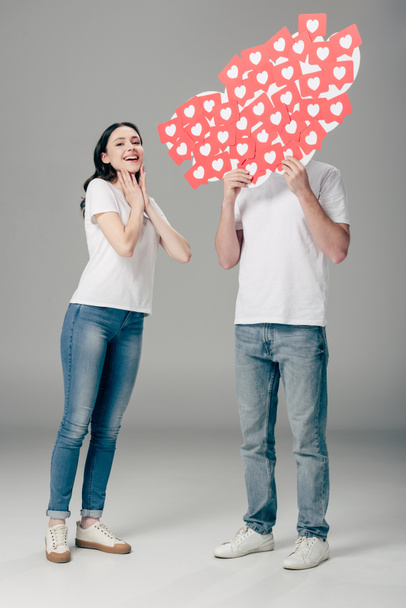 young man hiding face behind red paper cut cards with hearts symbol near excited girl on grey background - Photo, Image