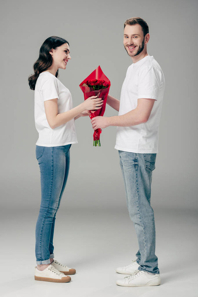 pohledný mladý muž, který se k hezké přítelkyni na šedém pozadí s kyticí červenými růží - Fotografie, Obrázek