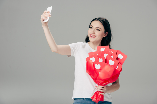 smiling girl holding bouquet of red paper cut cards with hearts symbol and taking selfie with smartphone isolated on grey - Foto, afbeelding