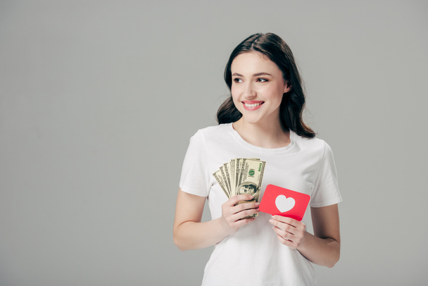 ドル紙幣と赤い紙切りカードを持つ美しい笑顔の少女は、灰色に隔離されたハートのシンボルを持つ - 写真・画像