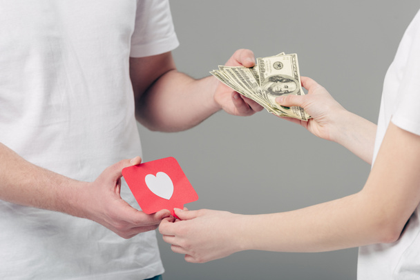 vista parcial del hombre que da billetes en dólares a la mujer que sostiene la tarjeta de papel rojo con el símbolo del corazón aislado en gris
 - Foto, imagen