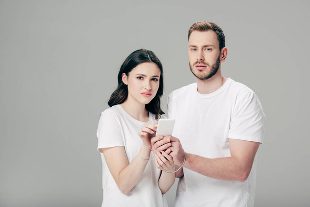 молодий чоловік і жінка в білих футболках з USB-кабелем навколо рук за допомогою смартфона ізольовані на сірому
 - Фото, зображення