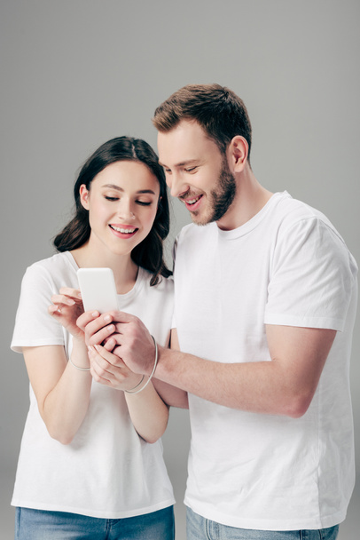 alegre hombre y mujer en camisetas blancas con cable USB alrededor de las manos usando teléfono inteligente aislado en gris
 - Foto, Imagen