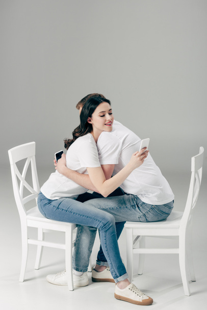 young couple embracing while sitting on white chairs and using smartphones on grey background - Foto, imagen