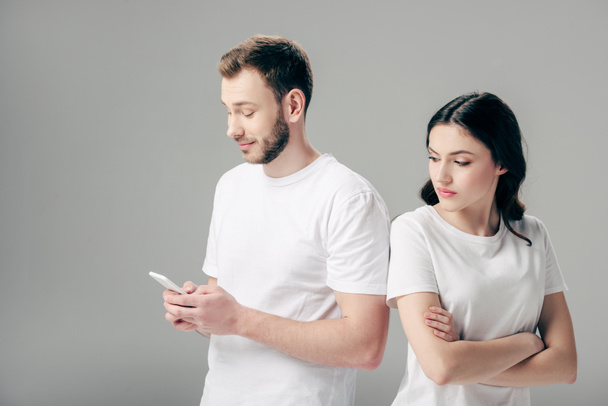 offended pretty girl standing with crossed hands near handsome boyfriend using smartphone isolated on grey - Photo, Image