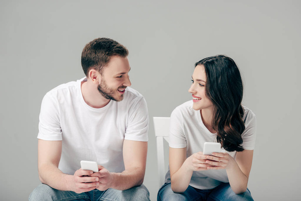 coppia sorridente guardarsi mentre si siede sulle sedie e utilizzando smartphone isolato su grigio
 - Foto, immagini