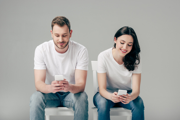 young couple in white t-shirts and blue jeans sitting on chairs and using smartphones on grey background - Photo, image