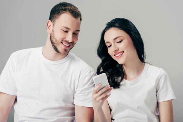 alegre joven hombre y mujer en camisetas blancas usando teléfono inteligente aislado en gris
 - Foto, imagen