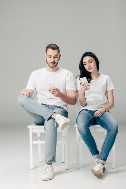 focused man and woman in white t-shirts and blue jeans sitting on chairs and using smartphones on grey background - Фото, изображение