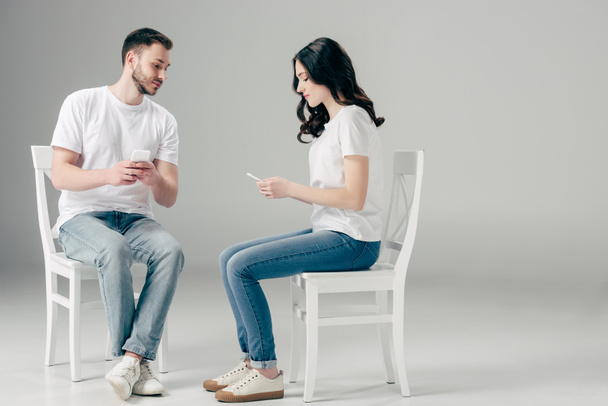 concentrated man and woman in white t-shirts and blue jeans sitting on chairs and using smartphones on grey background - Photo, Image