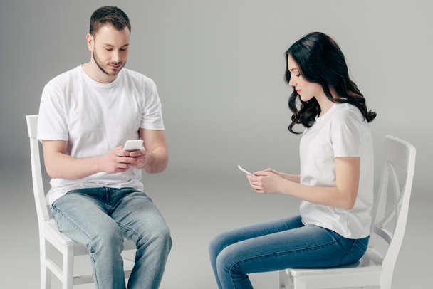 focused man and woman in white t-shirts and blue jeans sitting on chairs and using smartphones on grey background - Photo, Image