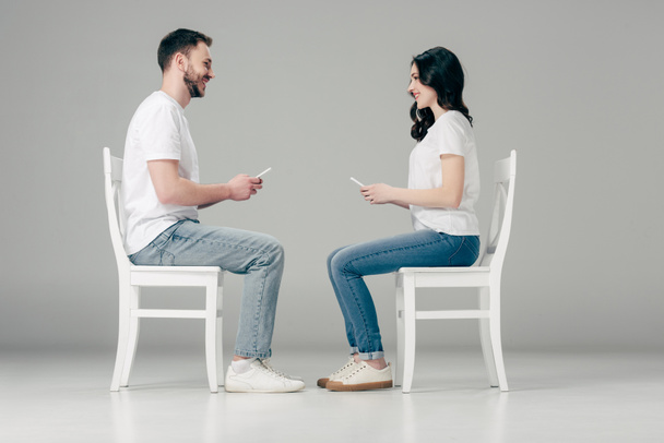 side view of smiling man and woman in white t-shirts and blue jeans sitting on chairs with smartphones and looking at each other on grey background - Photo, Image
