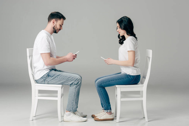 side view of concentrated man and woman in white t-shirts and blue jeans using smartphones while sitting on chairs on grey background  - Zdjęcie, obraz