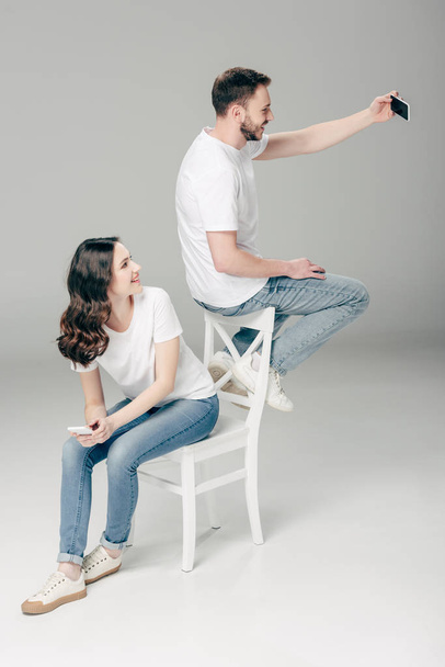 glimlachend meisje op zoek zittend op stoel en kijken naar vrolijke man het nemen van selfie met smartphone terwijl zittend op rug van stoel op grijze achtergrond - Foto, afbeelding