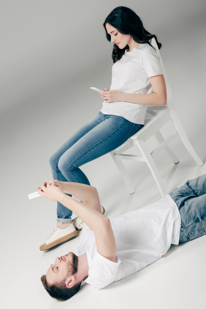 hübsche junge Frau sitzt auf Stuhl und benutzt Smartphone in der Nähe schöner Mann auf dem Boden liegend und macht Selfie auf grauem Hintergrund - Foto, Bild
