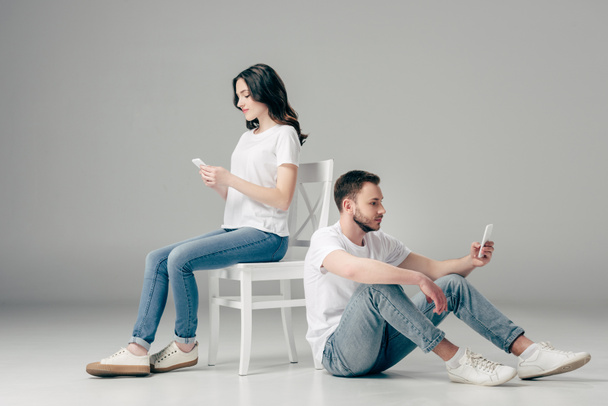 concentrated woman using smartphone near focused man sitting on floor with smartphone on grey background - Photo, Image