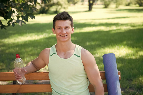 Towards healthier lifestyle. Man smiling face with yoga mat and water bottle sit on bench in park. Join outdoors yoga practice. Athlete with yoga equipment relaxing in park. Man chose yoga outdoors - Фото, изображение