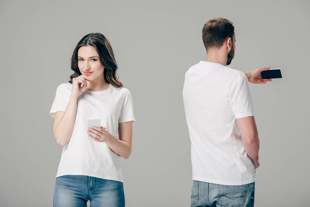 usmívající se atraktivní dívka v bílém tričku, stojící nedaleko mladého muže, který si bere telefon s smartphone izolovanými na šedé - Fotografie, Obrázek