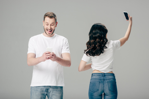 opgewonden jonge man met behulp van smartphone terwijl staande in de buurt meisje het nemen van selfie geïsoleerd op grijs  - Foto, afbeelding