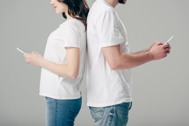 cropped view of cheerful man and woman in white t-shirts standing back to back and using smartphones isolated on grey - Photo, Image