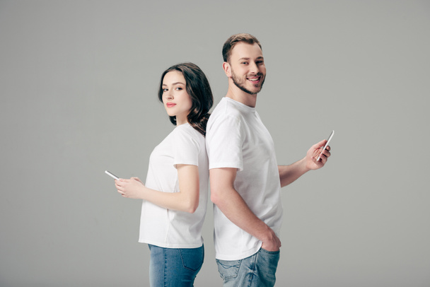 mladý usmívající se muž a žena v bílých tričkách stojí zády k sobě a pomocí chytrých telefonů izolovaných na šedé - Fotografie, Obrázek