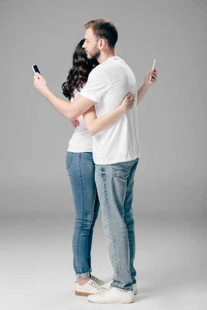young couple in white t-shirts and blue jeans embracing while using smartphones on grey background - Фото, изображение