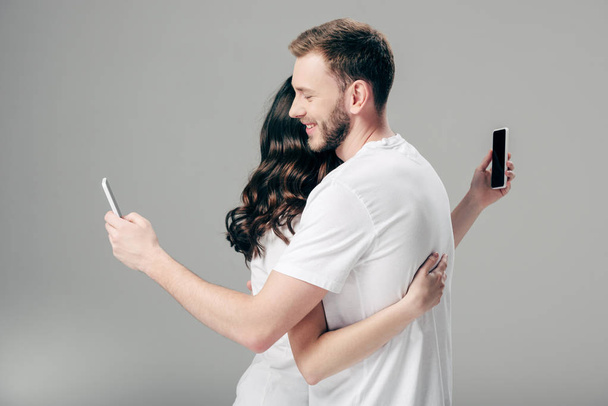 young couple in white t-shirts hugging while using smartphones on grey background  - Photo, Image