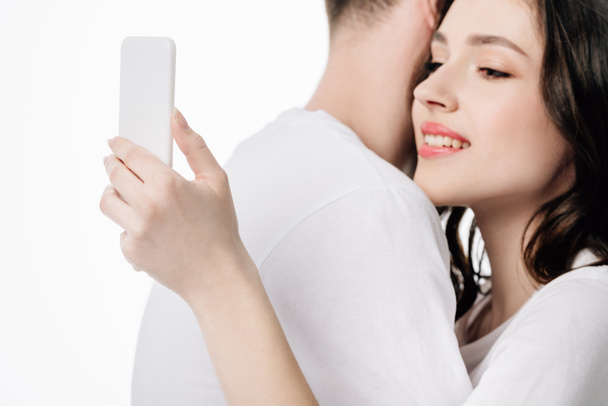 ελκυστική χαμογελαστή κορίτσι χρησιμοποιώντας smartphone, ενώ αγκάλιασμα γκόμενος απομονωμένος σε λευκό - Φωτογραφία, εικόνα