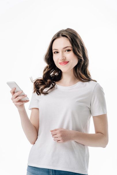 hübsches lächelndes Mädchen in weißem T-Shirt, Smartphone in der Hand und Kamera isoliert auf weiß blickend - Foto, Bild