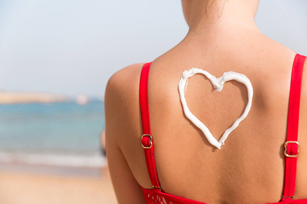 Beautiful girl in red swimsuit has sunblock in shape of heart at the back at the beach - Photo, Image