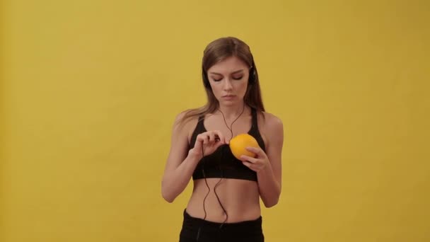 Sporty girl connecting headphones to orange and listens to music. Studio, yellow background. - Video, Çekim