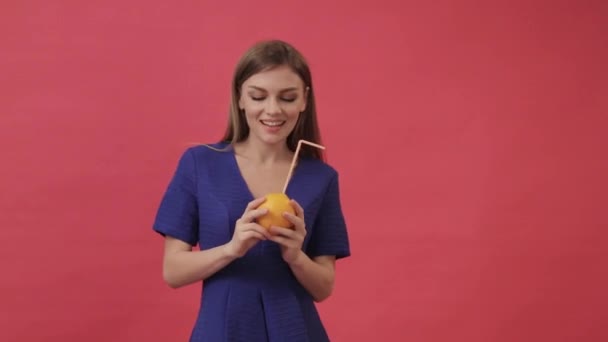 Beautiful girl in a dress drinking orange juice with a cocktail straw and dancing. Studio, purple background - Imágenes, Vídeo