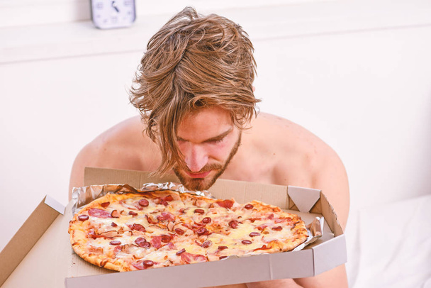 Food delivery service. Portrait of lazy muscular man eating pizza while laying on a bed at home. Man bearded handsome guy eating cheesy food for breakfast in bed. Pizza bed. - Фото, изображение