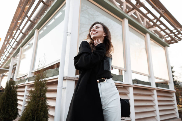 Счастливая молодая женщина в модной черной футболке в роскошном длинном пальто в винтажных джинсах с милой улыбкой позирует на улице рядом с летним кафе. Городская радостная девушка модель наслаждается прогулкой
. - Фото, изображение