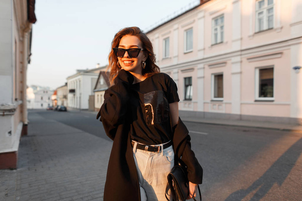 Happy young hipster woman in trendy clothes in stylish sunglasses walks around the city and enjoys the bright orange spring sunset. Beautiful joyful girl travel on a sunny day outdoor. Modern style - Foto, Imagen