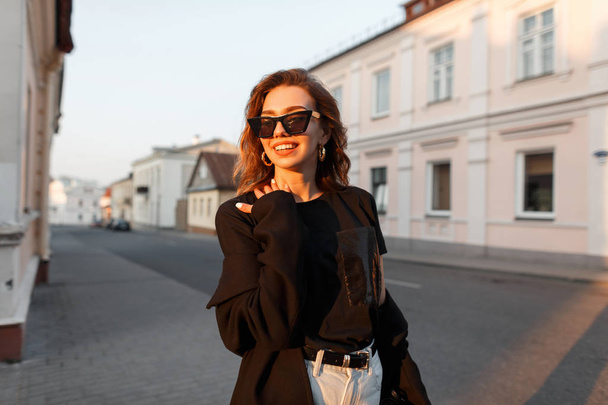 Χαρούμενη νεαρή χίππη γυναίκα σε σκοτεινά γυαλιά ηλίου με ένα χαμόγελο σε ένα κομψό παλτό σε ένα μπλουζάκι σε λευκό τζιν ταξιδεύει στο δρόμο κοντά σε vintage κτήρια σε μια φωτεινή ηλιόλουστη μέρα. Θετικό κορίτσι σε εξωτερικούς χώρους. - Φωτογραφία, εικόνα