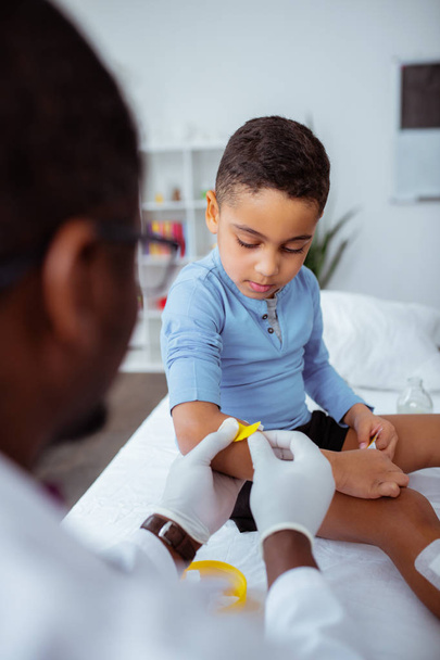 Dark-skinned doctor putting bright plaster on arm for little boy - Фото, зображення