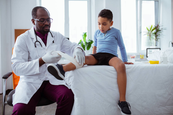 Dark-skinned pediatrician putting bandage on leg of little boy - Photo, Image