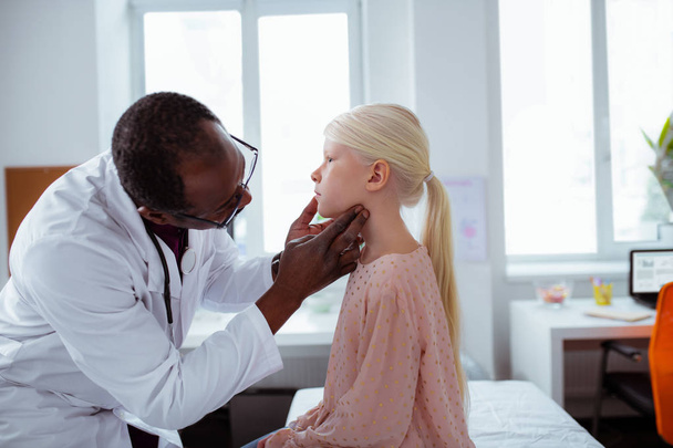Dark-skinned doctor wearing white coat looking at throat of girl - Foto, Imagen