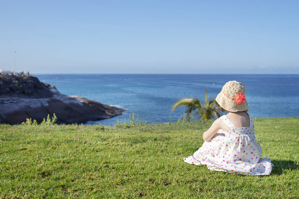 明るい光の夏のドレスと大きな花を持つわら帽子を身に着けている愛らしい白人の小さな女の子は、きちんとカットされた草の上に休んで、彼女の前の海岸の風景に向かって見て - 写真・画像