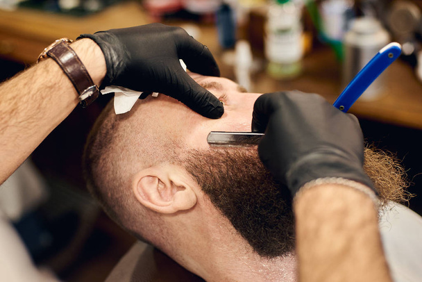 Klient liegend im Stuhl für die Rasur durch Friseurhände in Handschuhen mit scharfer Klinge im bärtigen Gesicht - Foto, Bild